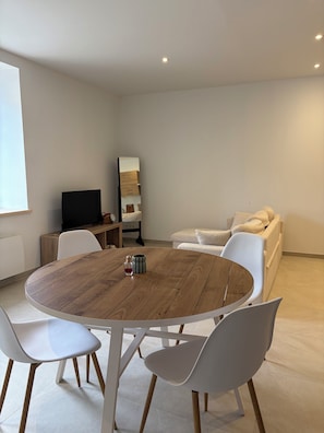 A modern living room with a round wooden table, white chairs, a sofa, and a flat-screen TV. - Cosy house with garden, quiet, near beach & port (paimpol)