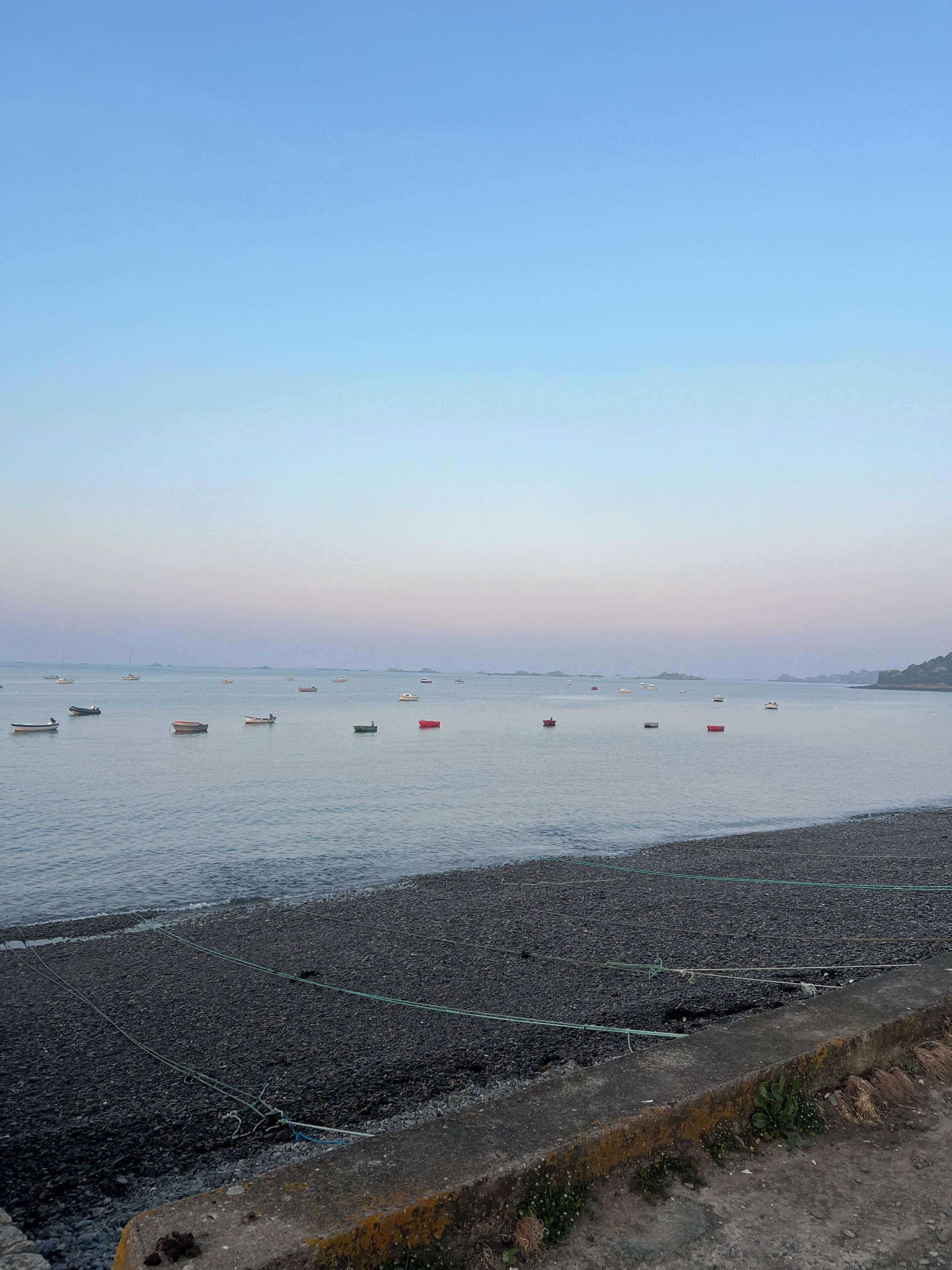 A pebbly beach with boats in the water and a clear sky.