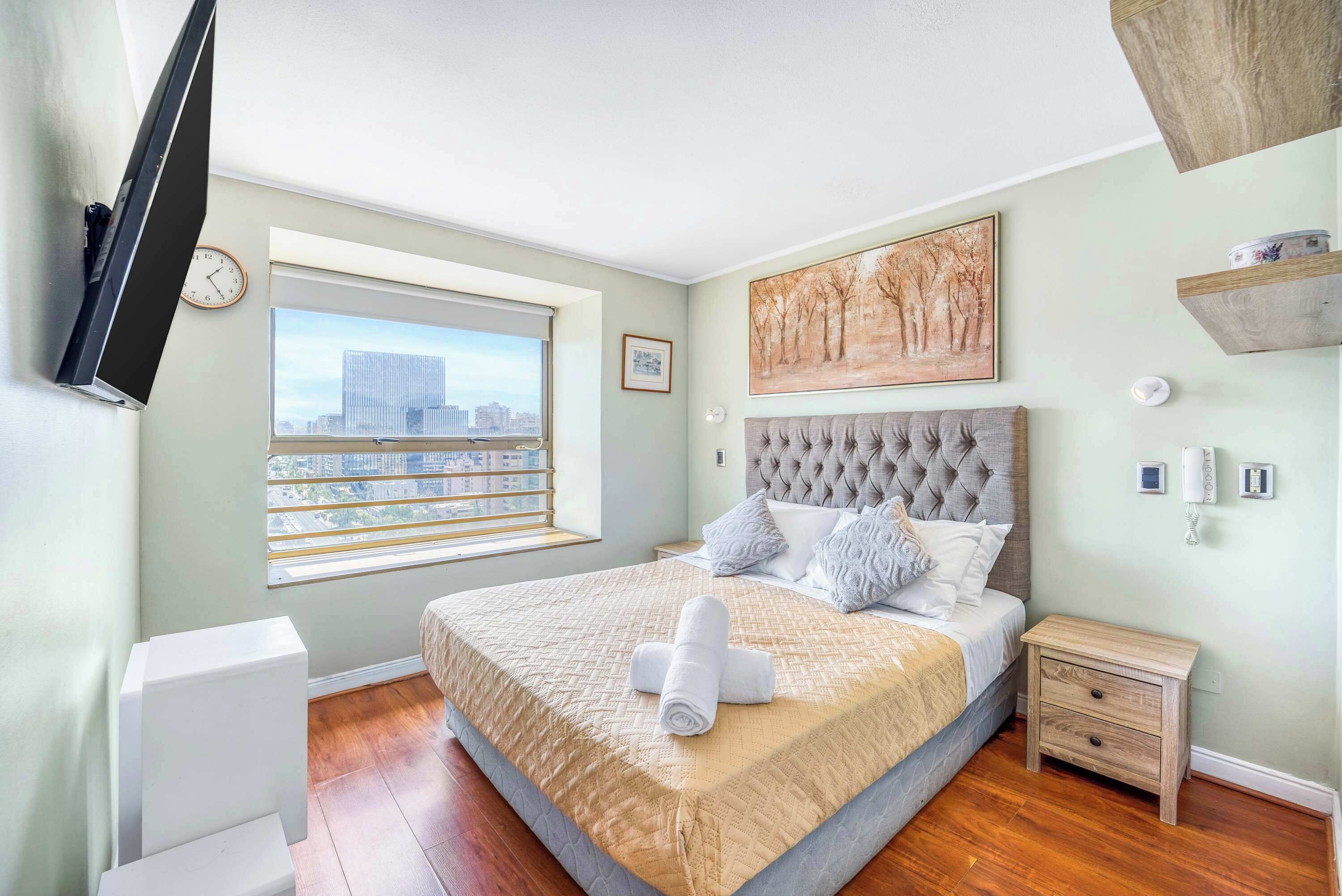 A bedroom with a large bed, a window with a city view, a mounted TV, and a painting on the wall.