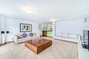 Ein modernes Wohnzimmer mit Couchtisch aus Holz, zwei weißen Sofas, einem gerahmten Kunstwerk und einer Küche im Hintergrund.