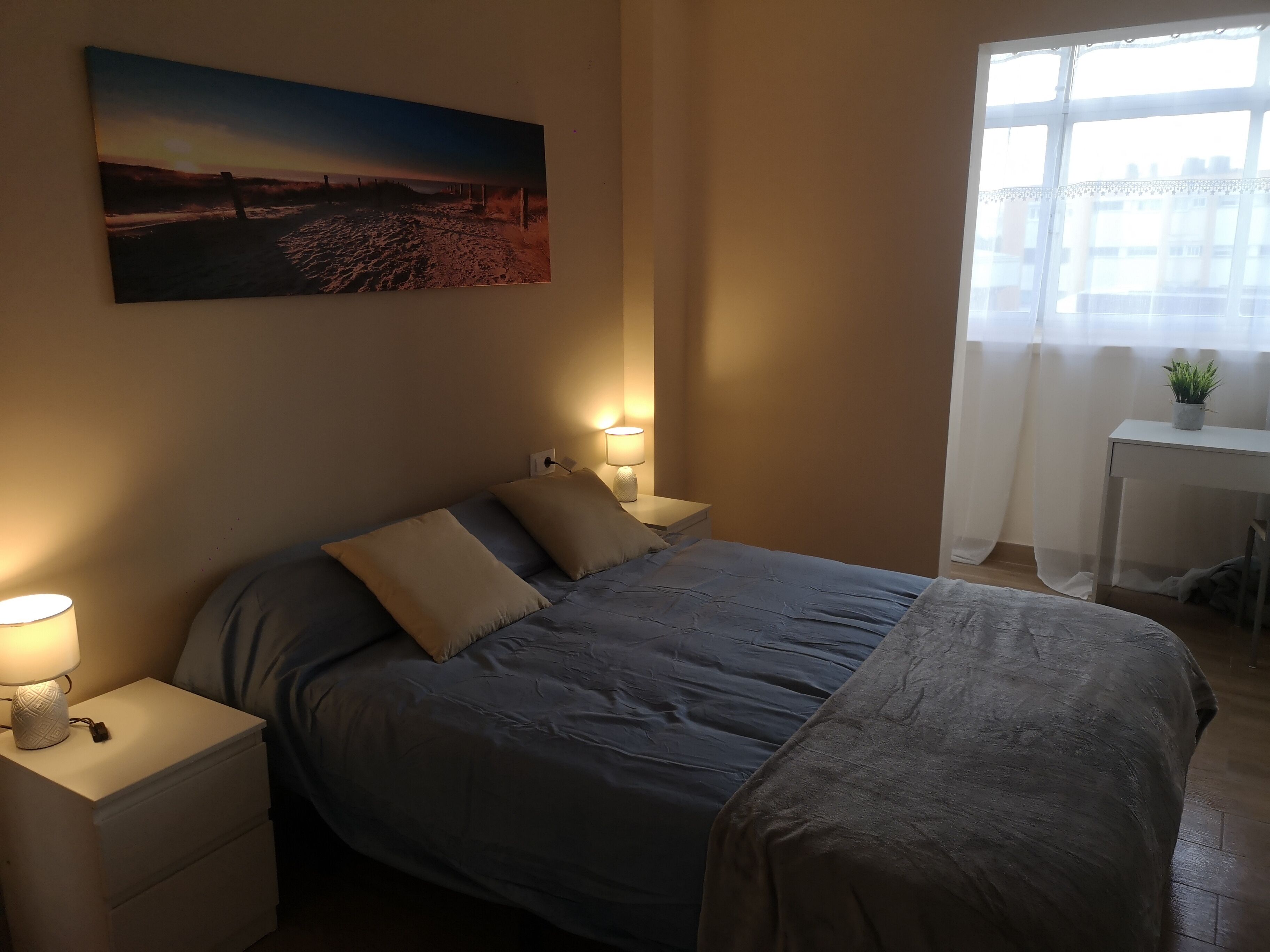 Un dormitorio con cama, mesitas de noche, lámpara, un cuadro, ventana y un escritorio con una planta.