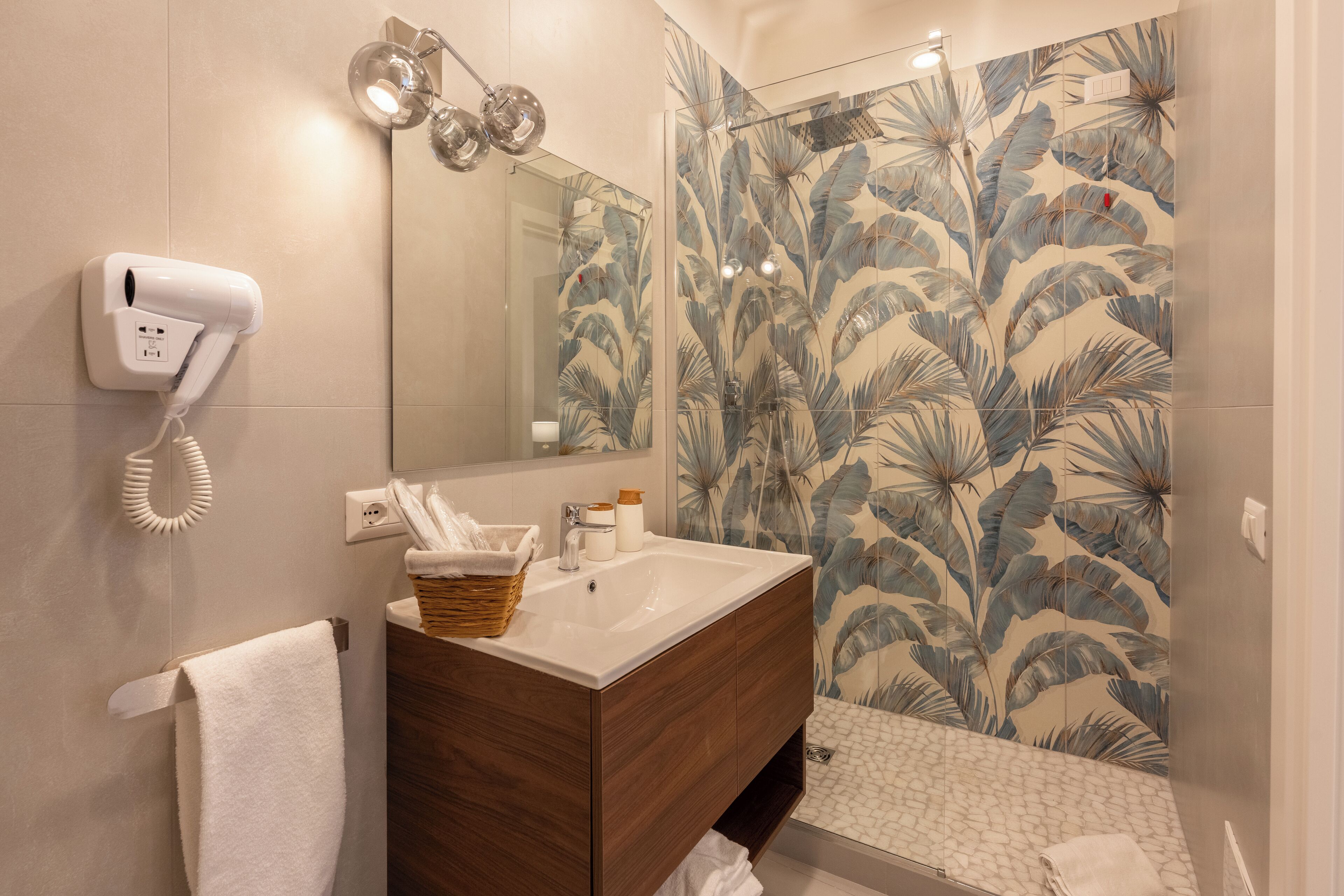A bathroom with a wooden vanity, a white sink, a mirror, a hairdryer, and a shower with a leaf-patterned tile wall.