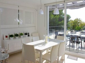 Ein moderner Essbereich mit einem weißen Tisch und Stühlen, einem Tisch mit Glasplatte und Blick ins Freie durch große Fenster.