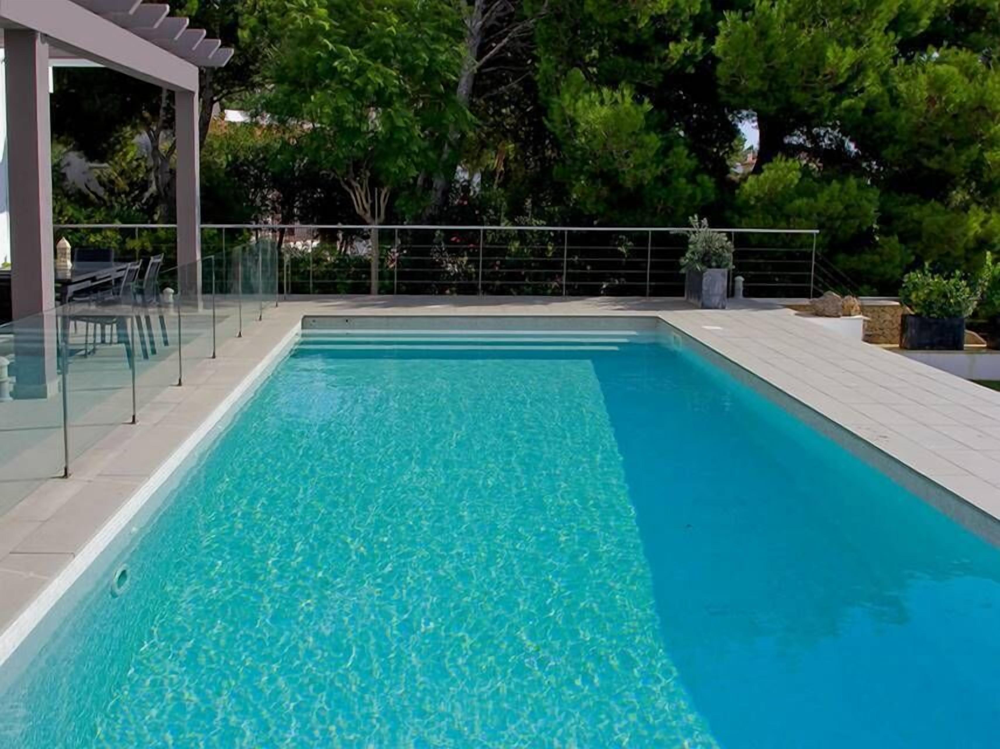 Una piscina rectangular amb aigua transparent de color blau, envoltada per un solàrium amb rajoles i una barana de vidre.