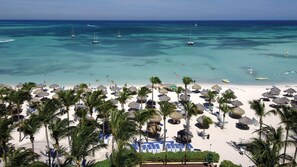 A beach with thatched umbrellas and palm trees, a sandy shore, and boats in the water. - Marriott's Aruba Surf Club | 2BR/2BA Oceanside (Noord)