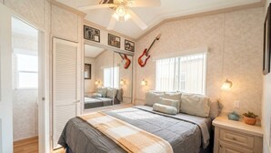 Une chambre à coucher avec un lit, un ventilateur de plafond, une guitare, un miroir et une fenêtre.