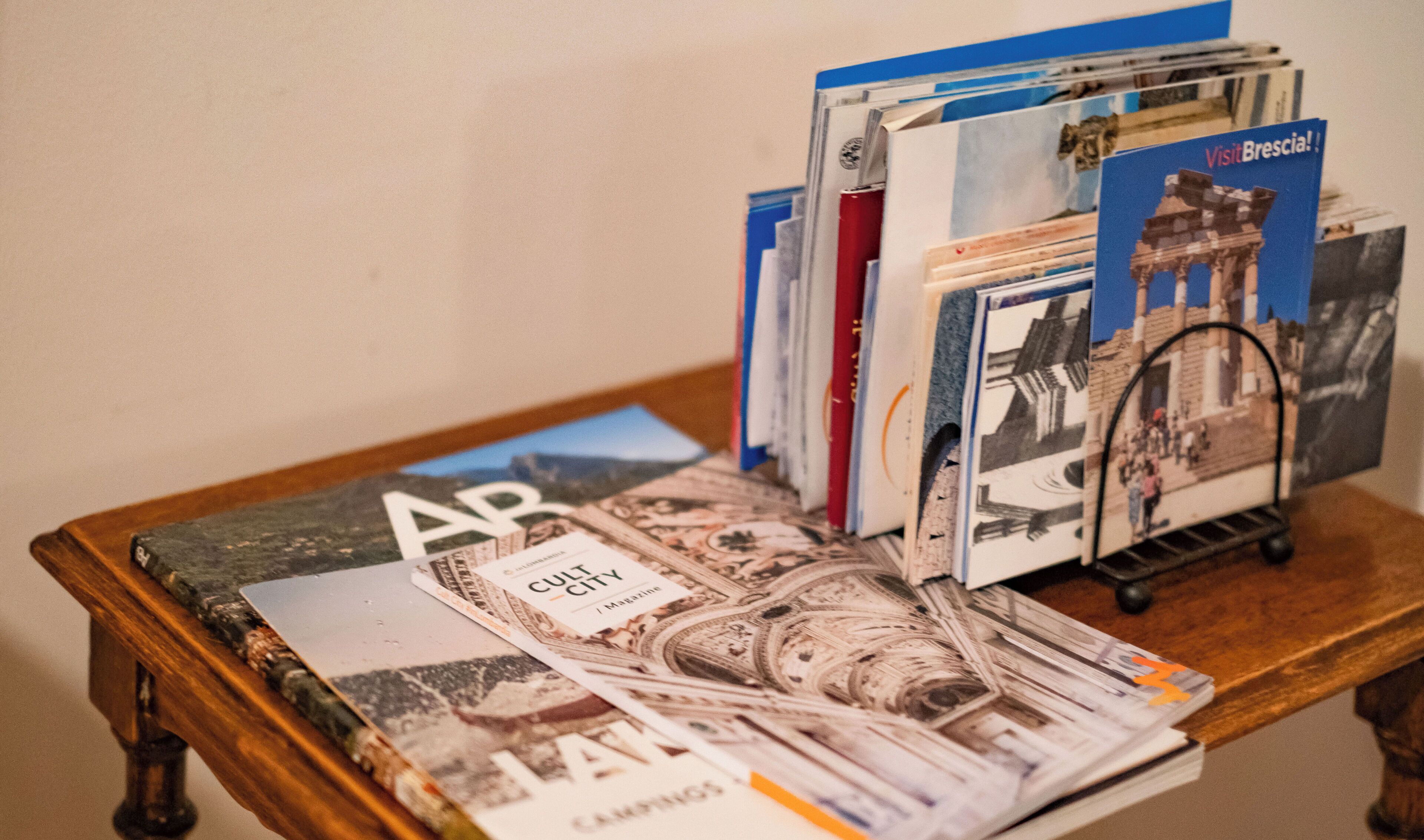 Une table en bois sur laquelle sont empilés des magazines et des brochures, dont une intitulée « Visitez Brescia ! »