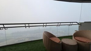 A balcony with wicker chairs and a glass railing overlooking a cityscape. - spira.staycation (Noida)