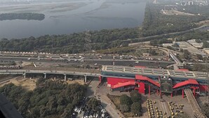 An aerial view of a coastal area with a large body of water, a bridge, and a red building with multiple sections. - spira.staycation (Noida)