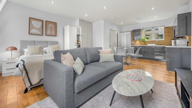 Een moderne woonkamer met een grijze bank, een klein rond tafeltje en een keuken op de achtergrond.