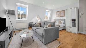 Een moderne woonkamer met een grijze bank, een salontafel en een televisie. Er is een raam met uitzicht op gebouwen, een radiator en een kledingkast.