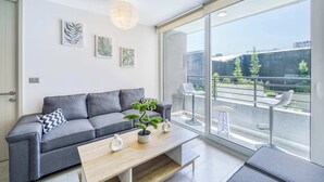 A modern living room with a grey sofa, a wooden coffee table, and a potted plant. There are framed artworks on the wall and a balcony with outdoor seating. - Live Ñuñoa: conexión L1/L3 y vida urbana (Ñuñoa)