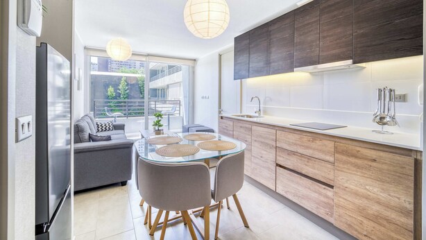 A modern kitchen with wooden cabinets, a glass dining table, and a view of the outdoors. - Live Ñuñoa: conexión L1/L3 y vida urbana (Ñuñoa)