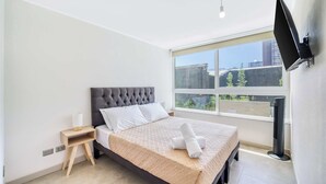 A modern bedroom with a large bed, a nightstand, a flat-screen TV mounted on the wall, and a window with a city view. - Live Ñuñoa: conexión L1/L3 y vida urbana (Ñuñoa)