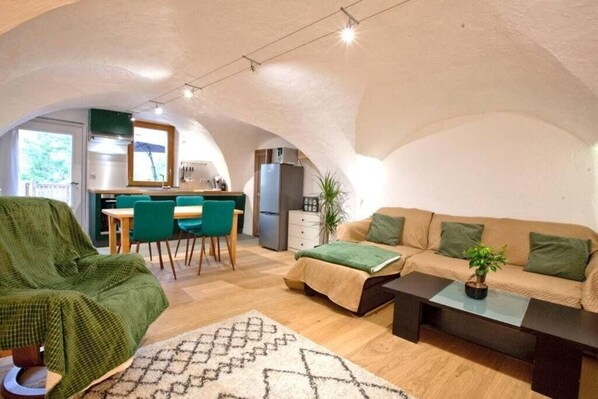 Un salon accueillant avec un canapé, une table basse et un fauteuil vert. On aperçoit une cuisine à l’arrière-plan, avec une table à manger et des chaises.