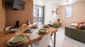 A modern living room with a dining area, featuring a wooden dining table set with plates and glasses, a sofa with cushions, a TV mounted on the wall, and a window with a view of a snowy landscape. - Bright apartment for 7 people in Les Deux Alpes (Les Deux Alpes)
