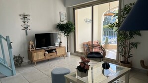 Een moderne woonkamer met een flatscreen televisie, een glazen salontafel, een rieten fauteuil en een potplant.