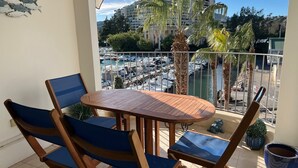 Een balkon met een houten tafel en blauw gestoffeerde stoelen, met uitzicht op een jachthaven vol boten en een hoog gebouw op de achtergrond.