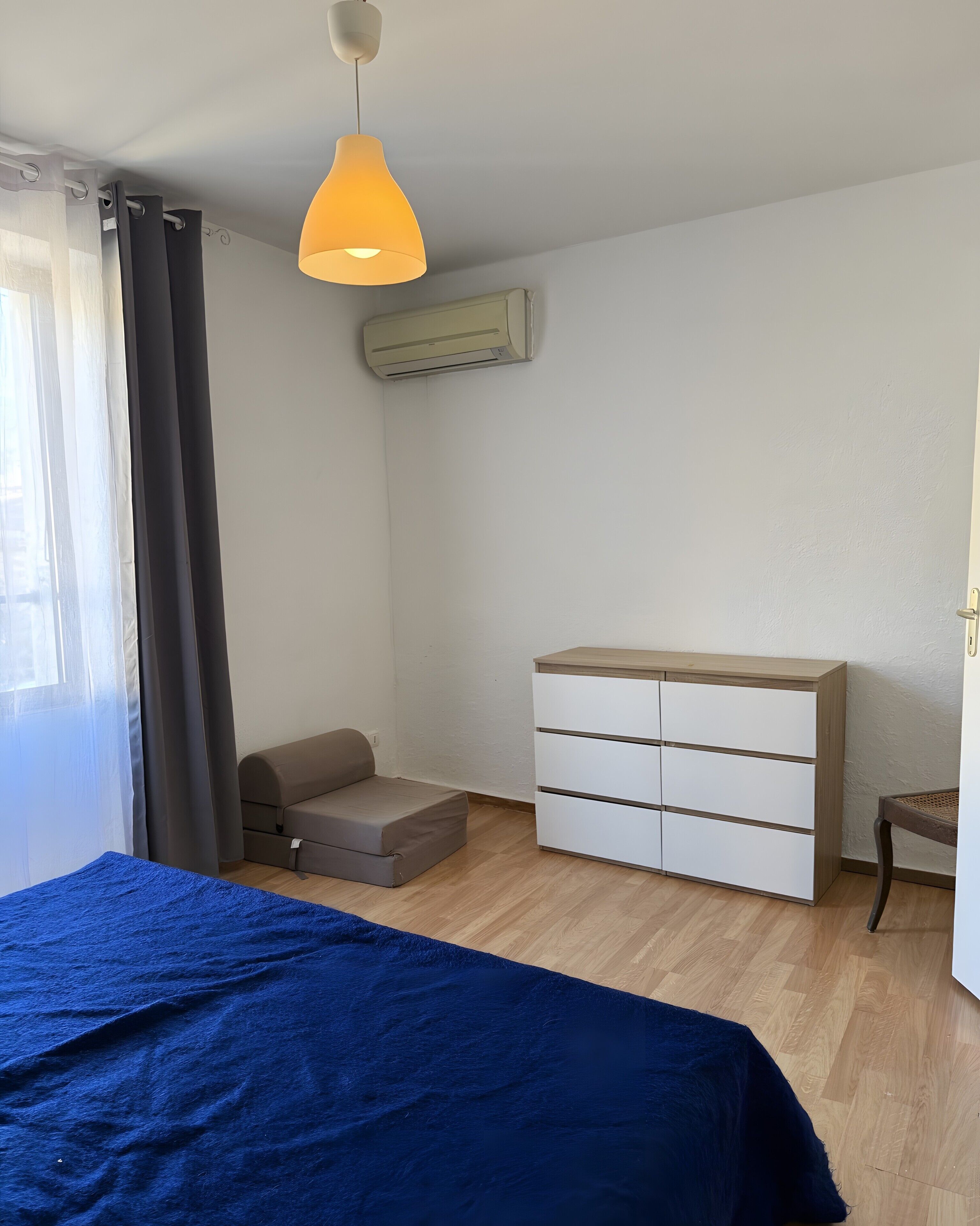 A bedroom with a bed, a dresser, a sofa, an air conditioning unit, and a hanging light fixture.