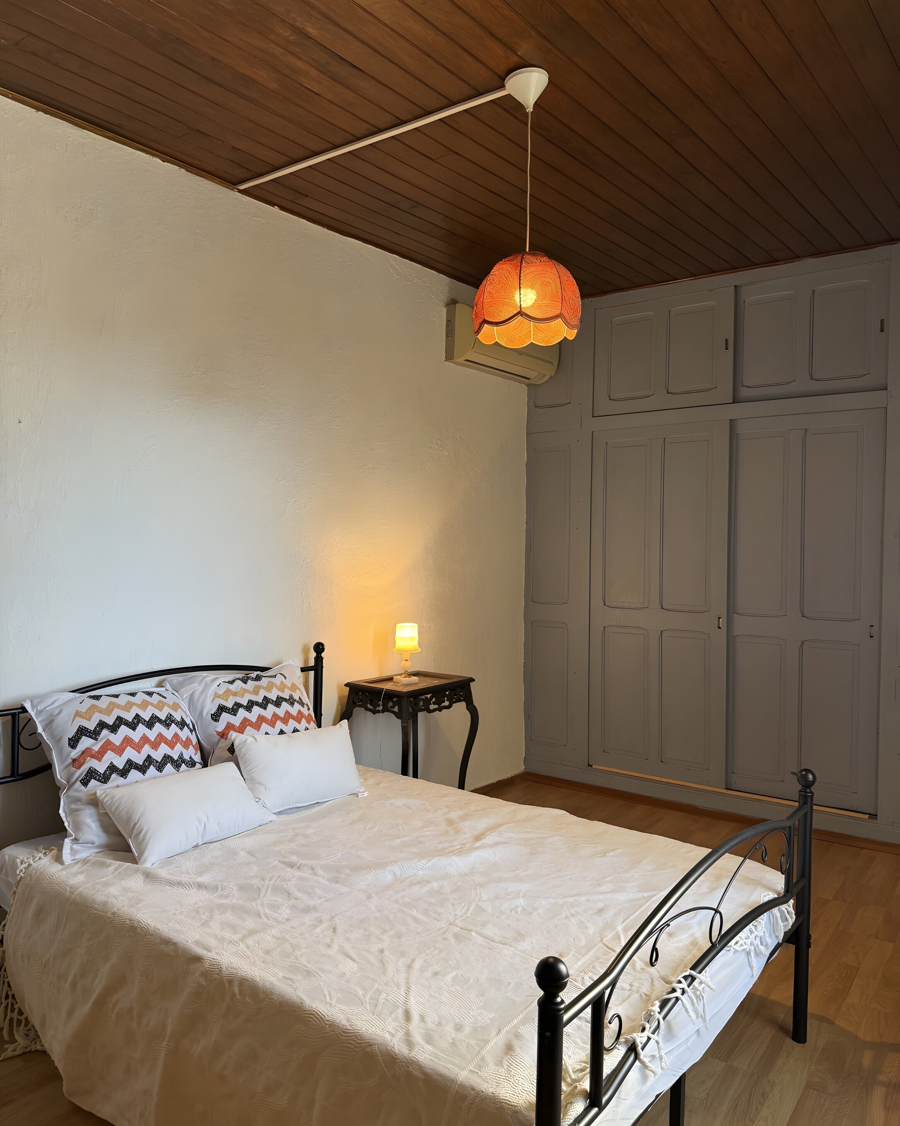 A bed with a white bedspread, two pillows, a bedside table with a lamp, and a hanging light fixture.
