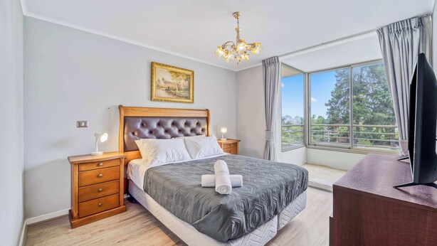Ein Schlafzimmer mit einem großen Bett, einem Nachttisch aus Holz, einem Schreibtisch, einer Chandelierleuchtre, einem Gemälde und einem großen Fenster mit Blick.