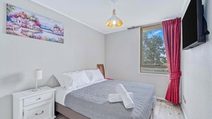 Ein Schlafzimmer mit Bett, Nachttisch, Lampe, Gemälde, Fenster und Vorhängen.