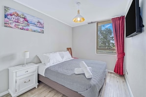 A bedroom with a bed, nightstand, lamp, painting, window, and curtains.