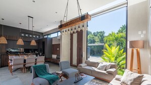 A modern living room with a sofa, armchairs, and a coffee table. There is a dining area with a table and chairs. The room has large windows with a view of greenery outside. - Design Villa · Tulum Country Club · PGA Golf (Gran Bah ía Príncipe)