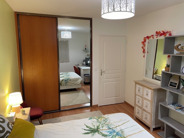 A bedroom with a bed, a desk, a chair, a wardrobe, a mirror, and a shelf with decorative items.