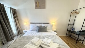 A neatly made bed with pillows and folded towels, a bedside table with a lamp, and a wall-mounted shelf with a hanging garment rack.