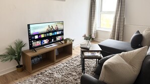 A living room with a flat-screen TV, a sofa, a coffee table, a plant, and a window with curtains.