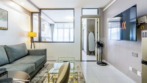 Ein modernes Hotelzimmer mit grauem Sofa, einem Glastisch, einem Flachbildfernseher, einem Standventilator und einem großen Fenster mit Blick auf den Flur und ein weiteres Zimmer.
