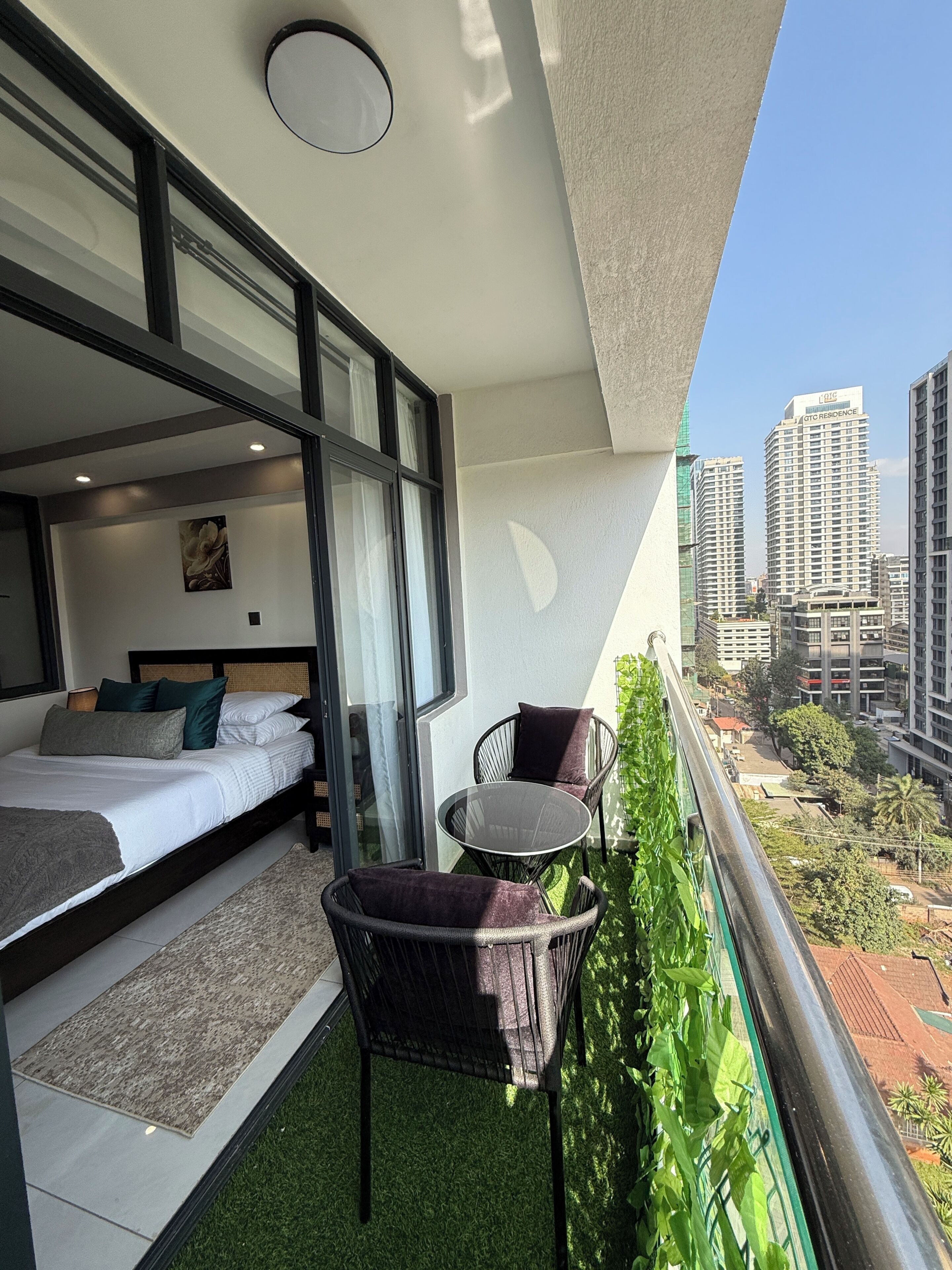 A balcony with a bed, a small table, and chairs, overlooking a cityscape with high-rise buildings.