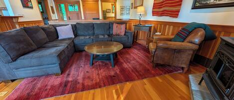 Ein Wohnzimmer mit einem Ecksofa, einem runden Couchtisch, einem Ledersessel, einem Kamin und einem roten Teppich.