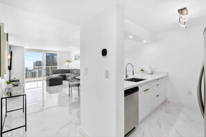 A modern kitchen with a built-in dishwasher, a dining area with a gray sofa, and a large window offering a city view. - Renovated Ocean-View Lux Condo with Balcony & Parking (Miami)