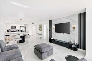 A modern living room with a flat-screen TV, a dining area, and a kitchen. - Renovated Ocean-View Lux Condo with Balcony & Parking (Miami)