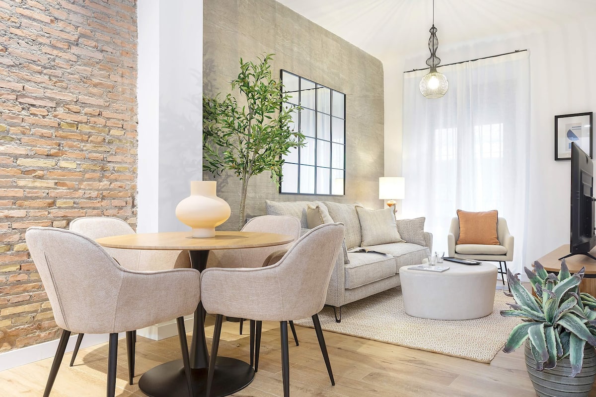 Un salón moderno con una mesa redonda, dos sillas, un sofá, una mesa auxiliar, una planta en maceta y un amplio ventanal con vistas.