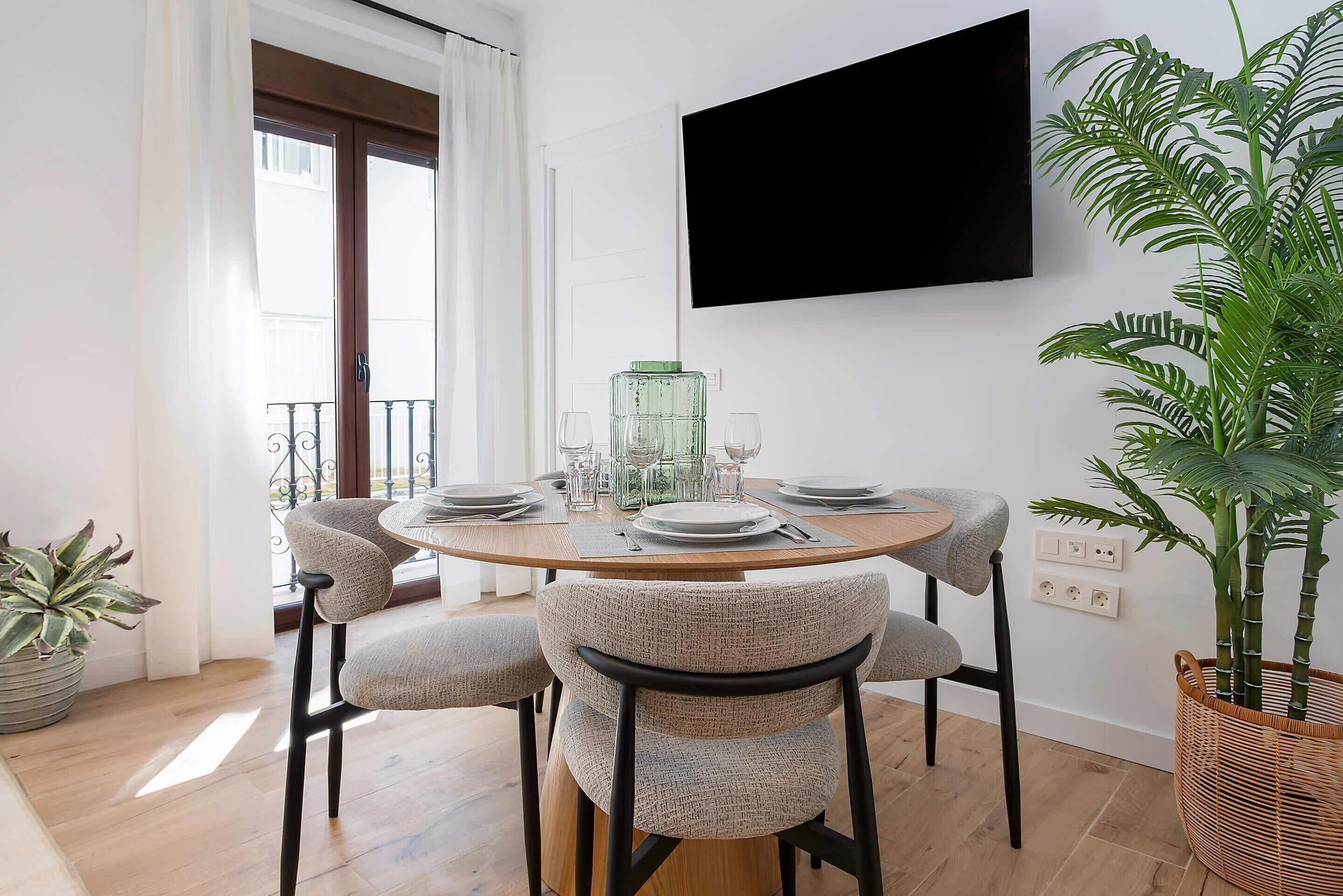 A dining area with a round table, four chairs, a TV mounted on the wall, and a potted plant.