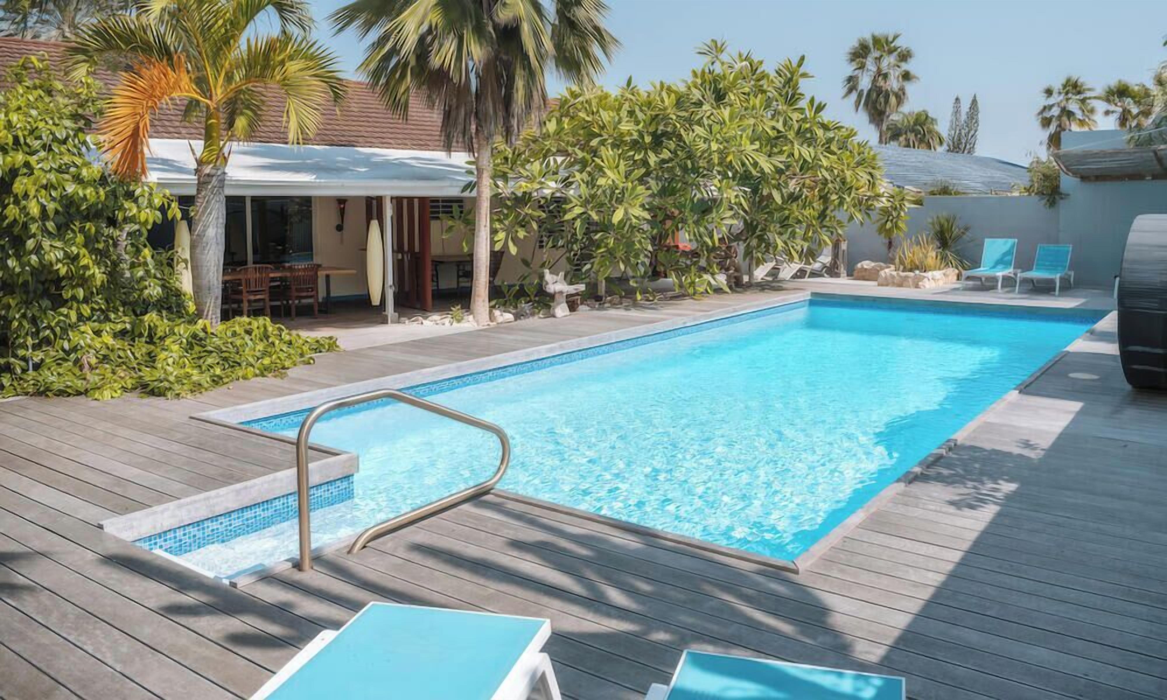 A swimming pool with a wooden deck, lounge chairs, and palm trees.