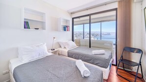 Une chambre d’hôtel moderne avec deux lits, une grande fenêtre donnant sur la ville, une bibliothèque et un fauteuil.