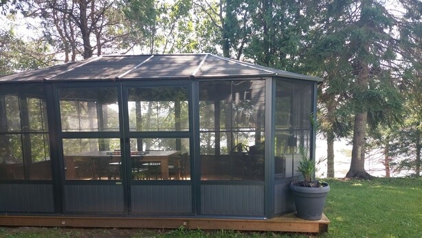 A covered patio with a wooden deck, glass walls, and a table with chairs. - La Maison Hacquard (4BR) - 35 min from Mont-Tremblant, Perfect for Families (St-Emile de Suffolk)