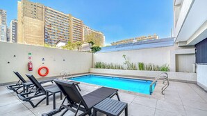A rooftop pool area with lounge chairs and a small table, surrounded by a high-rise building. - Central Isabel II with Pool, Wifi & AC near Metro Santa Isabel (Santiago)