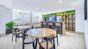 A modern indoor space with a round wooden table surrounded by chairs, a kitchen area with a brick backsplash, and shelves with plants. - Central Isabel II with Pool, Wifi & AC near Metro Santa Isabel (Santiago)