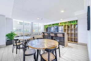 A modern indoor space with a round wooden table surrounded by chairs, a kitchen area with a brick backsplash, and shelves with plants. - Central Isabel II with Pool, Wifi & AC near Metro Santa Isabel (Santiago)