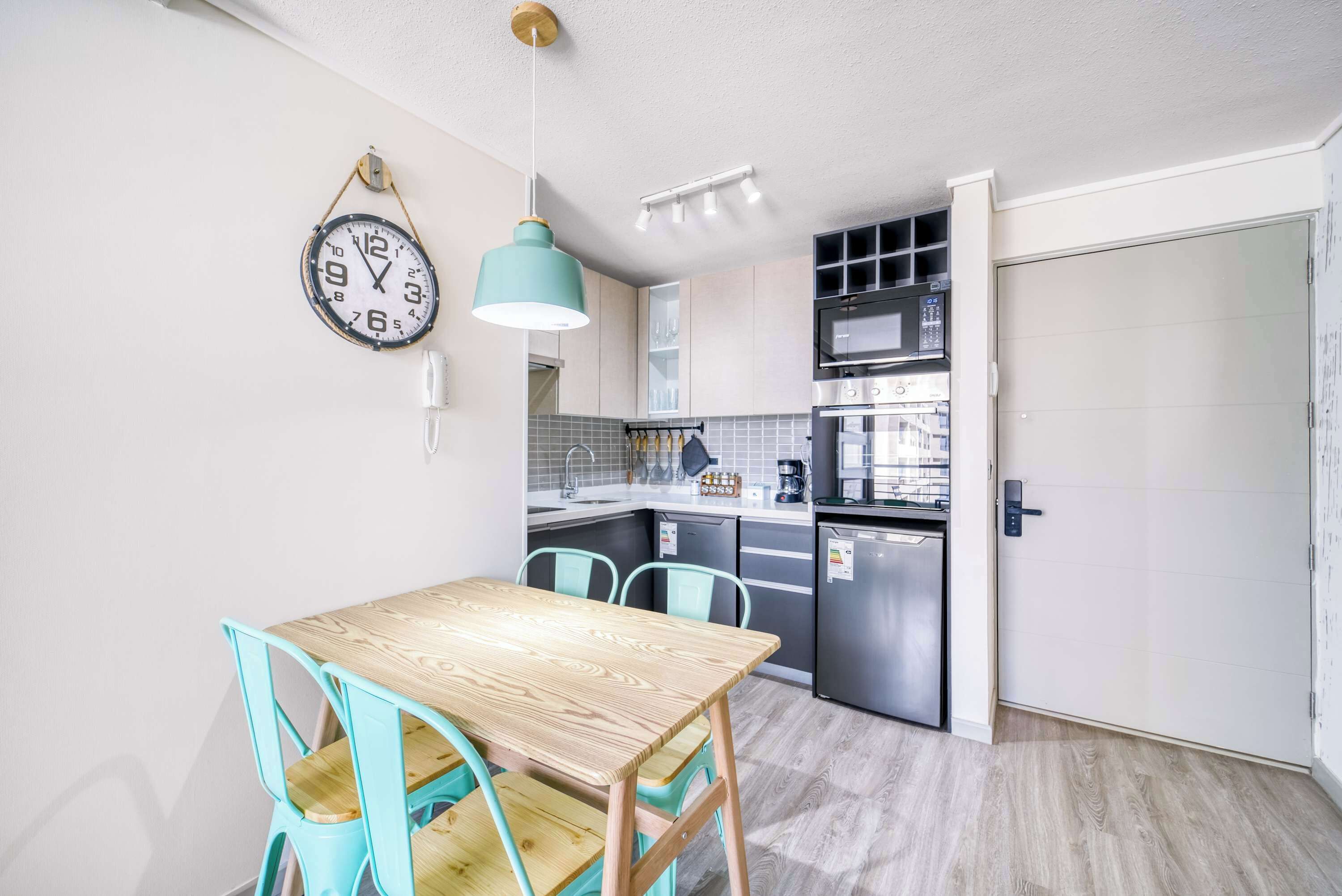 Une cuisine compacte avec une table en bois et des chaises turquoise, une horloge suspendue et des appareils électroménagers modernes.