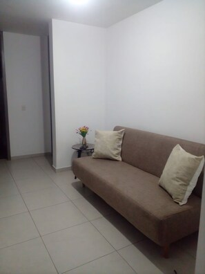 A living room with a brown sofa, two beige cushions, a small round table with a vase of flowers, and a white wall.