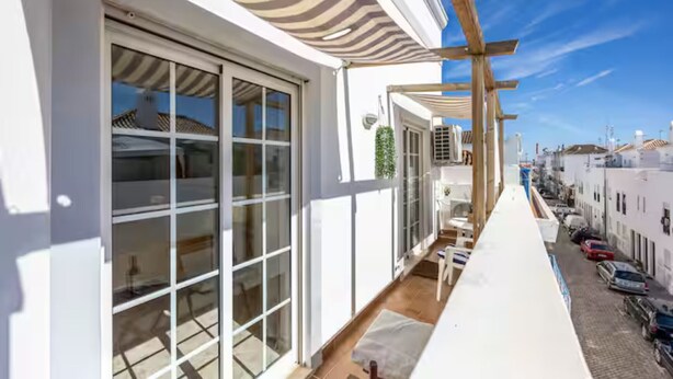 Ein Balkon mit Blick auf parkende Autos und Gebäude, eine Pergola mit gestreifter Markise und ein Fliesenboden.