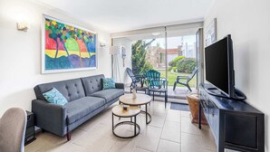 A modern living room with a grey sofa, a coffee table, a flat-screen TV, and a large window offering a view of the outdoors. - Central Reñaca W / Garden First Floor near Reñaca (Viña del Mar)