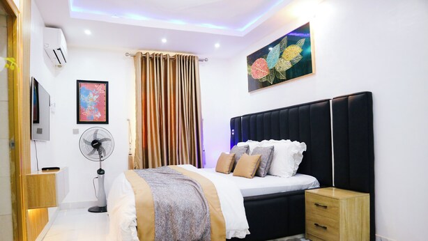 Une chambre moderne avec une tête de lit noire, un lit avec une literie blanche et beige, une table de chevet, une petite table de nuit en bois, un téléviseur fixé au mur, un ventilateur sur pied et une grande fenêtre avec des rideaux.