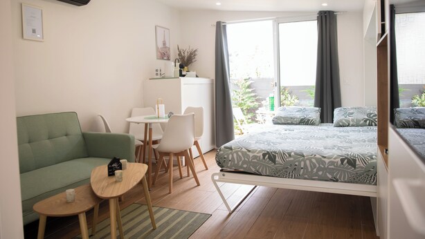 Una habitación moderna con un sofá verde, una mesita, una cama con ropa de cama estampada, un escritorio con sillas y una ventana con cortinas.
